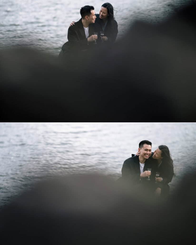 The couple laugh over champagne by the water, framed by a dark blurred foreground, on their proposal day at Mount Fuji.