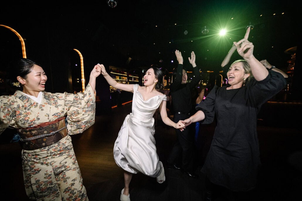 The bride dances hand in hand with a guest in a cream kimono and brocade obi as friends cheer under green light at the wedding dance party in Tokyo.
