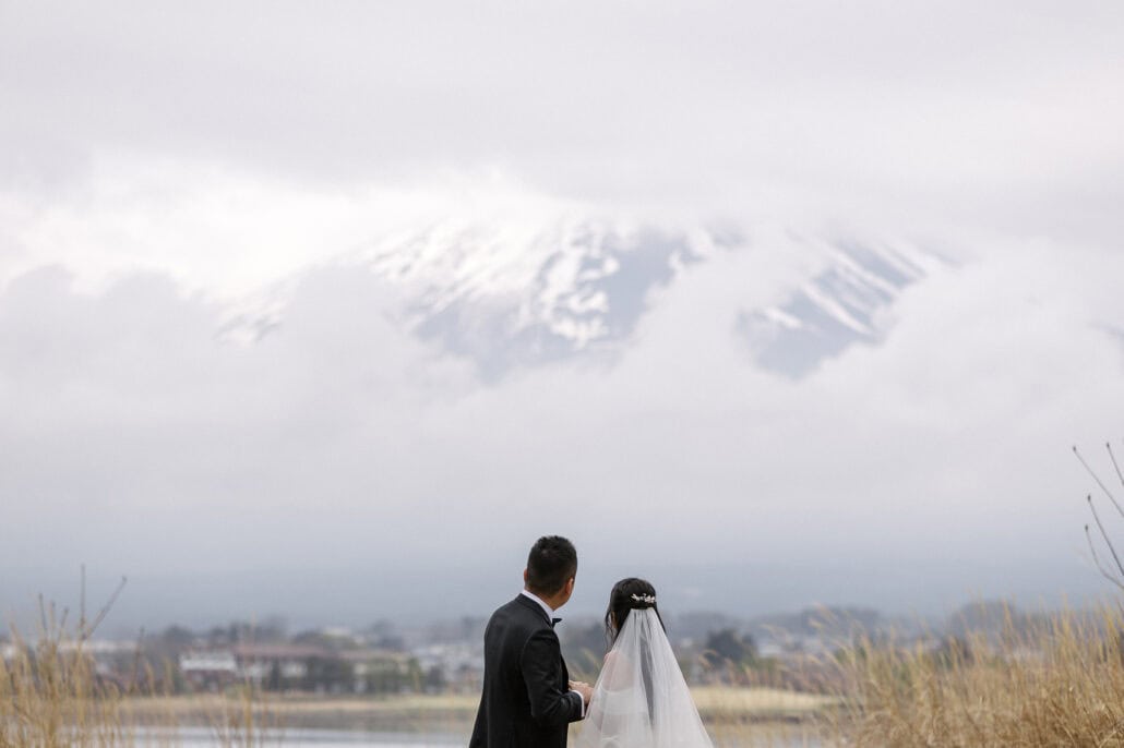 Seen from behind, the bride and groom stand in dry grass on the shore of Lake Kawaguchi, looking up at the snow on Mt Fuji in the cloud.