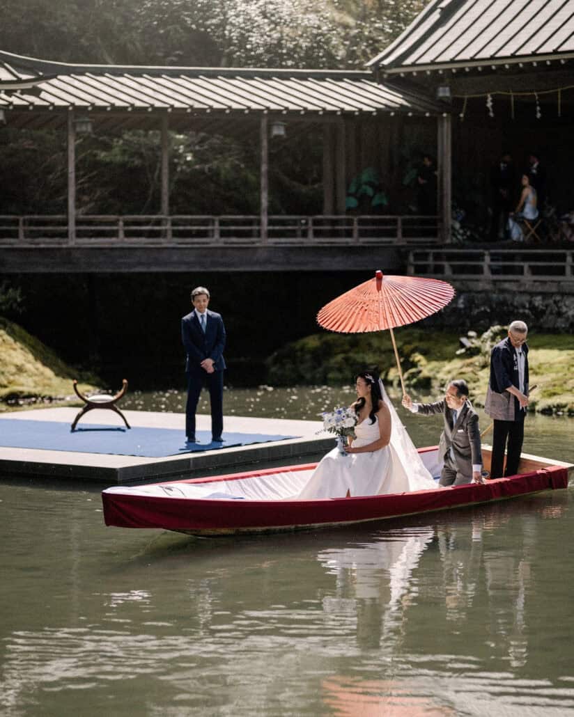 The bride arrives by boat under a red paper umbrella, her father waiting on the platform and guests watching from Asaba ryokan's Noh stage.