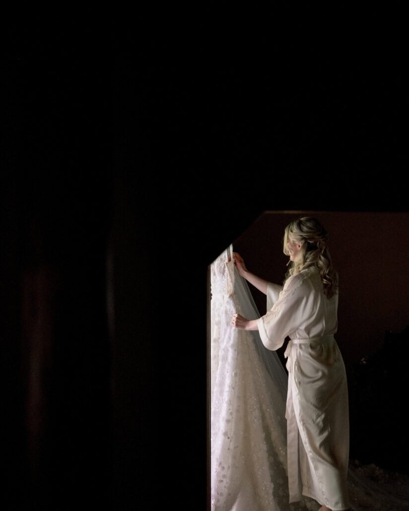 The bride in a satin robe reaches up to her sequinned wedding dress and veil hanging in a bright window, getting ready for an elopement at a ryokan.