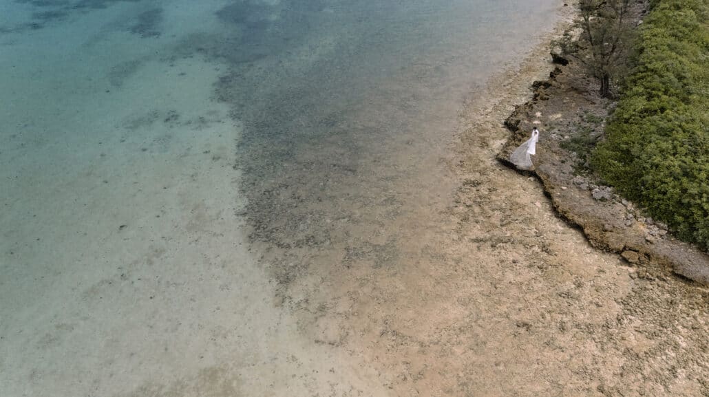 From above, the couple stand small on the reef rocks at Rosewood Miyakojima, her long veil spread over dark stone beside clear jade shallows.