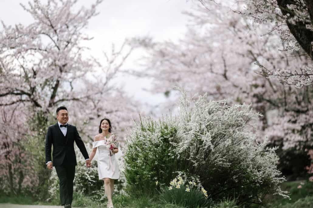 The bride and groom walk hand in hand toward the camera, smiling, past white spiraea and daffodils, her pink bouquet in hand.