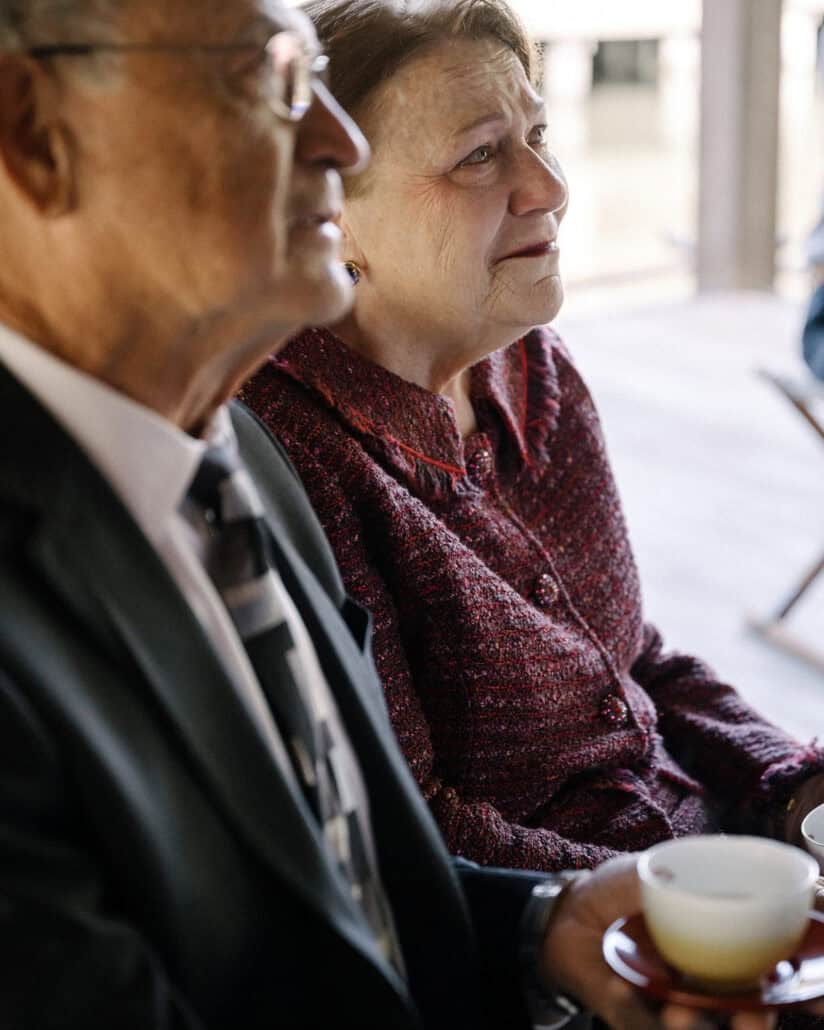 A father and mother sit side by side at the wedding tea ceremony at Asaba ryokan, his tea cup on a red lacquer saucer, her eyes wet.