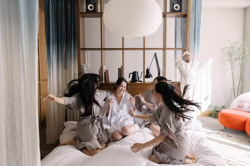 Three bridesmaids in taupe robes bounce on the bed around the laughing bride under a big paper lantern, the wedding dress hanging nearby at K5.