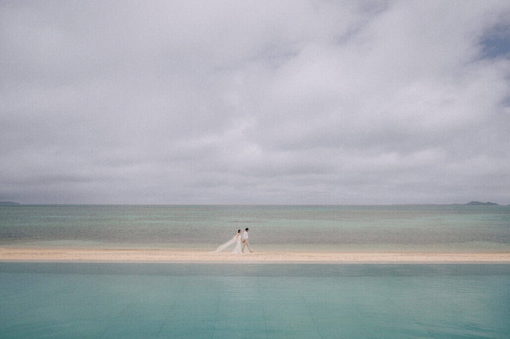 Beyond the edge of the infinity pool at Rosewood Miyakojima, the couple walk a strip of white beach under heavy cloud, her veil streaming out.