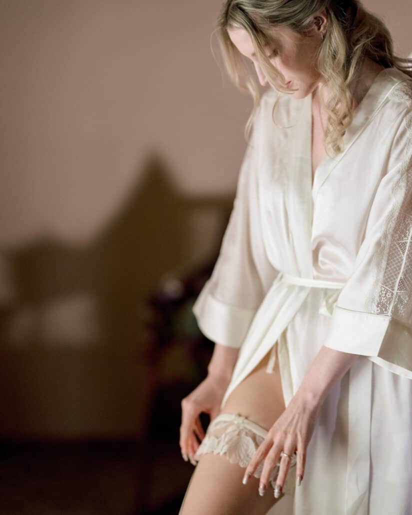 The bride in a satin robe slides a lace garter into place, getting ready in a softly lit room for her elopement in Japan.
