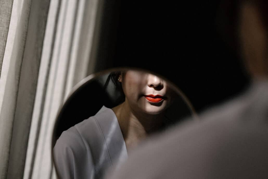 The bride's red lipstick and white robe caught in a round mirror, her face half in shadow, as she gets ready for her wedding in Tokyo.