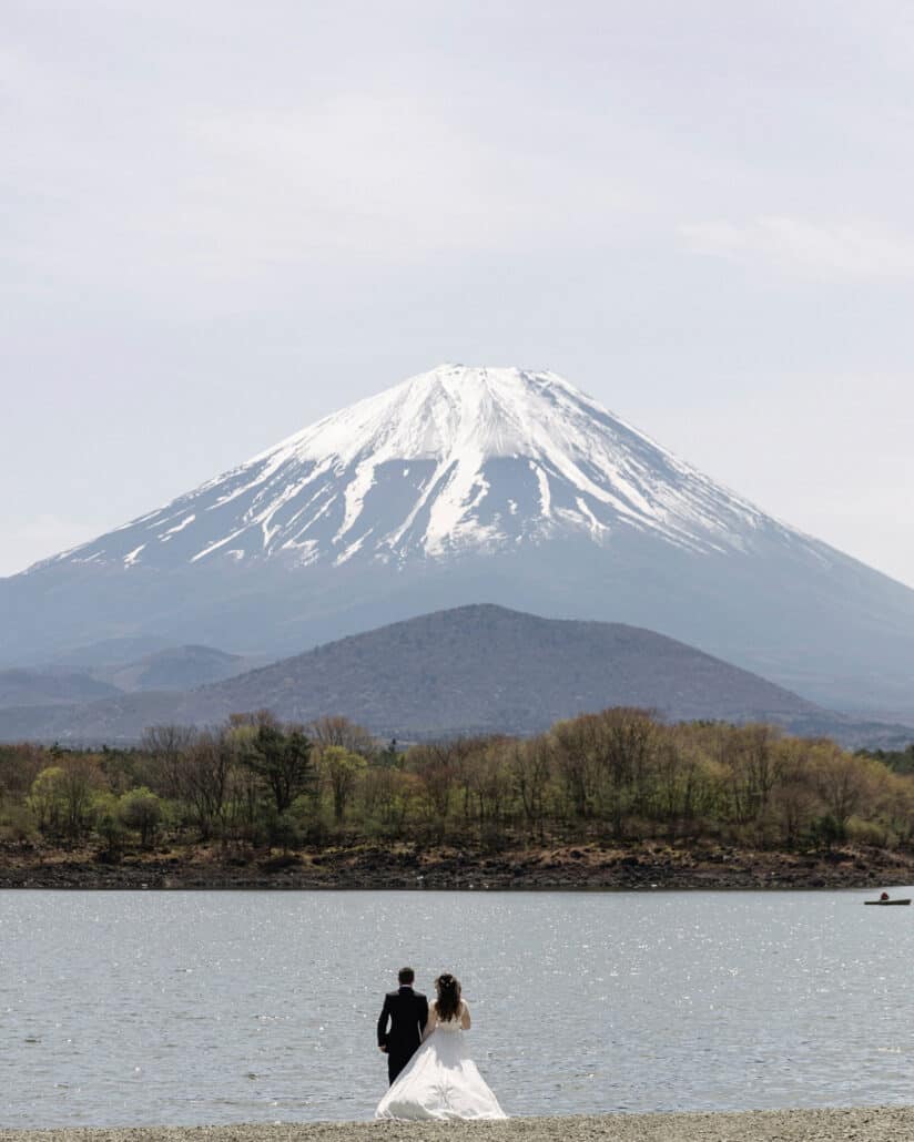 The couple stand at the water's edge of Lake Shoji, looking across to Mt Fuji and the smaller dark cone in front of it, a small boat out on the lake.
