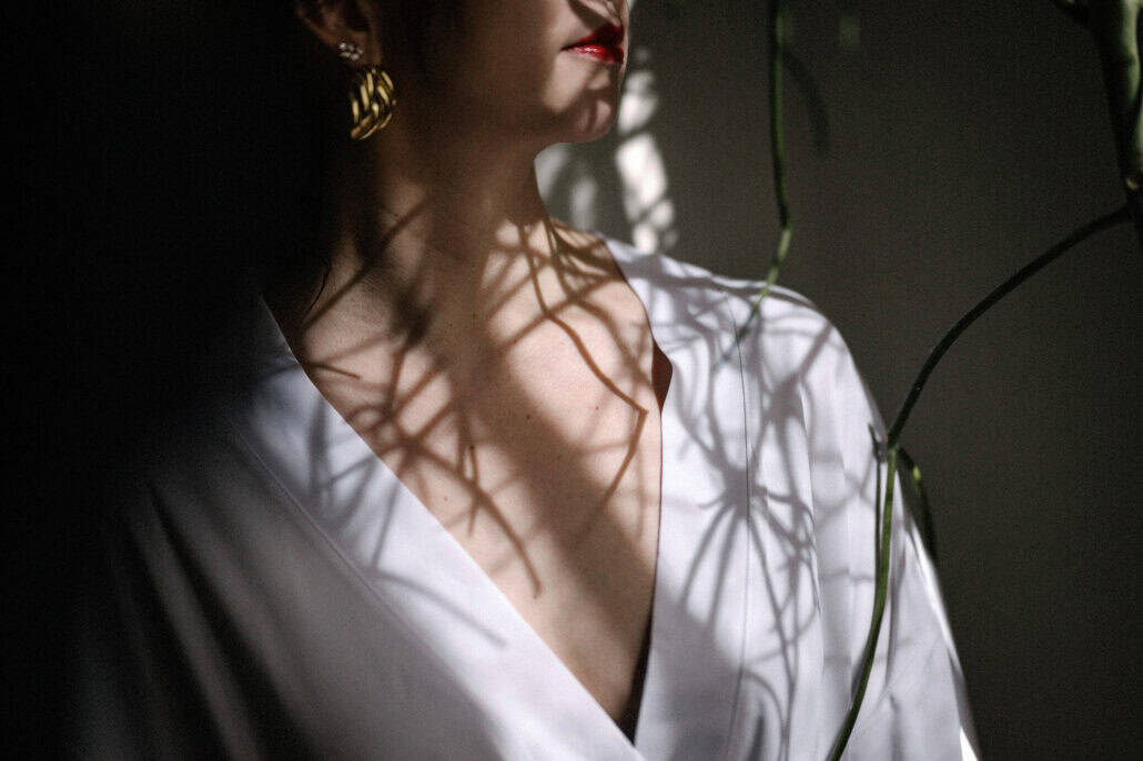 A close detail of the bride in her white robe, plant shadows across her collarbone, a gold statement earring and red lips on her wedding morning.
