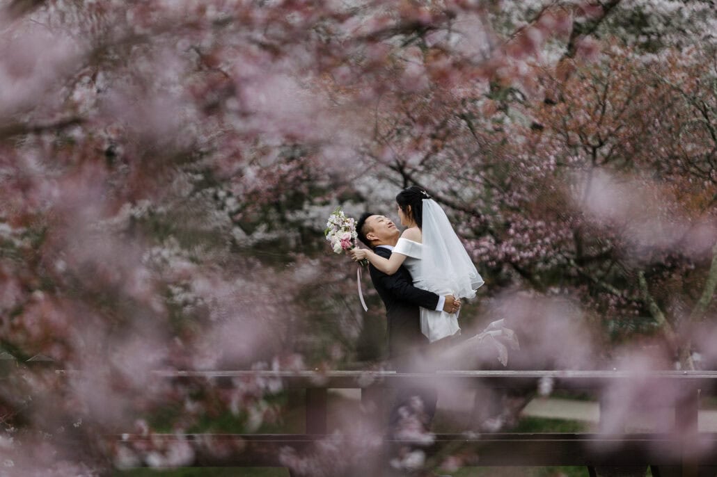 The groom lifts the bride on a wooden footbridge, her bouquet raised and heels kicked up, seen through a haze of pink cherry blossom.