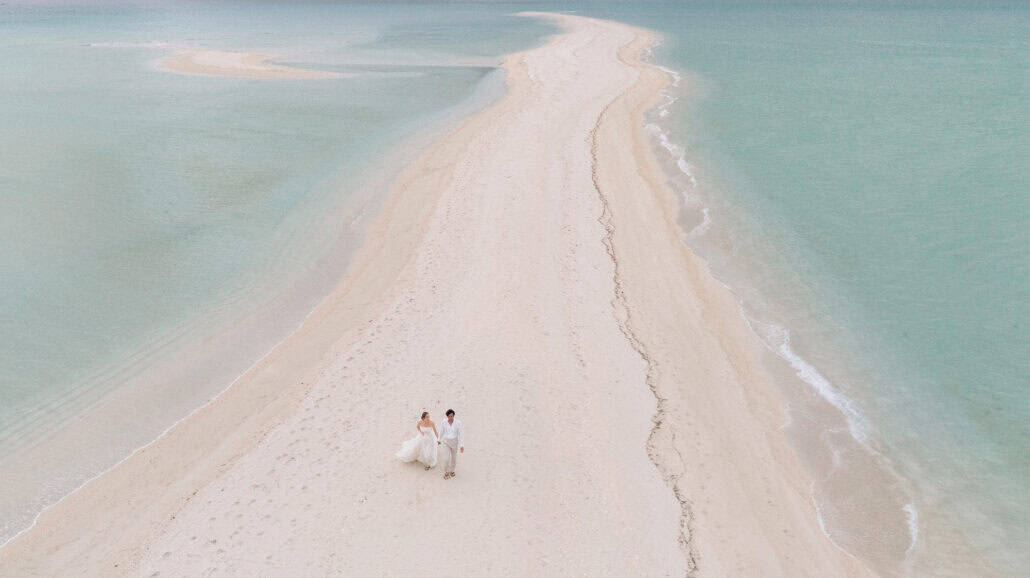 A long white sandbar curves away through turquoise shallows off Miyako Island, the bride in a flowing gown walking beside the groom.