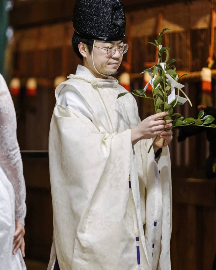A Shinto priest in white robes and a black hat carries the tamagushi, a sakaki branch with white paper, at the couple's shrine blessing in Japan.