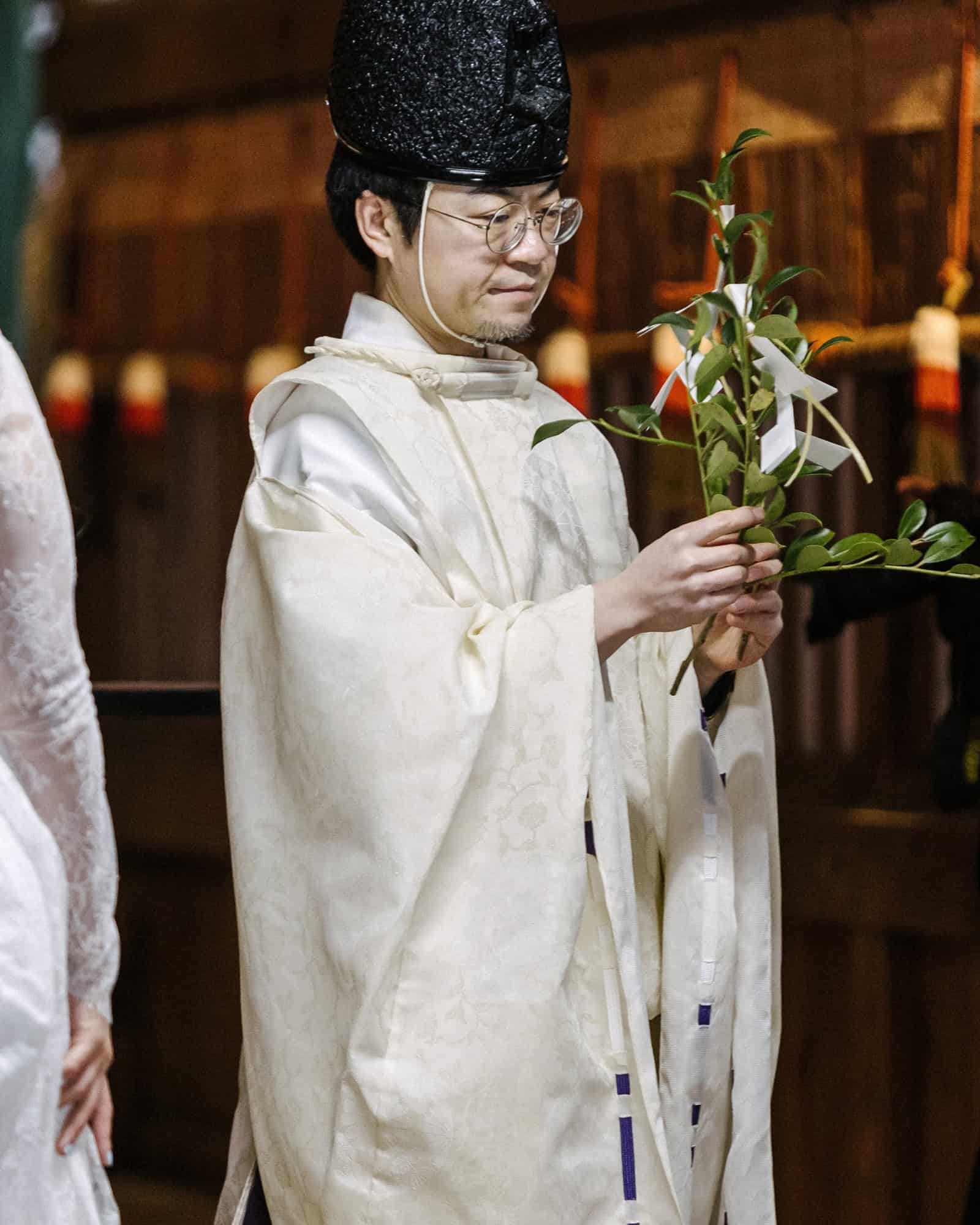 A Shinto priest in white robes and a black hat carries a tamagushi, a sakaki branch tied with white paper, for a shrine wedding blessing in Japan.