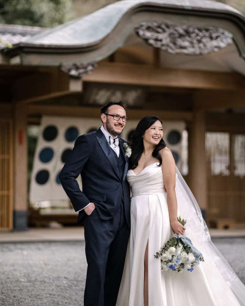 The bride and groom smile at the entrance of Asaba ryokan, its round blue windows and carved curved roof behind them, her bouquet tied in blue.