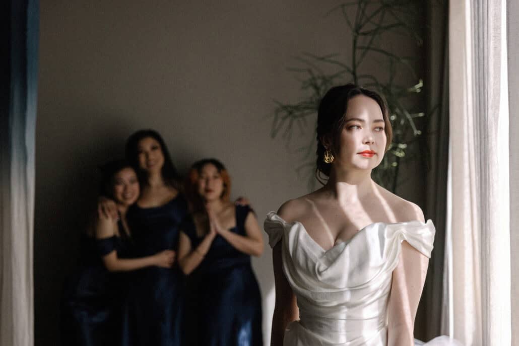The bride in her satin gown stands in window light while three bridesmaids in navy wait out of focus behind her at K5 in Kabutocho.