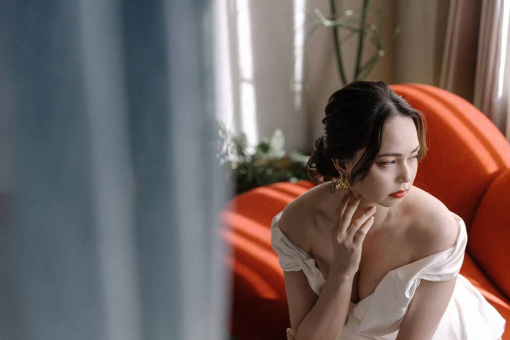 The bride in an off-shoulder satin gown sits on a red sofa with one hand at her neck, looking toward the window past a blurred curtain.