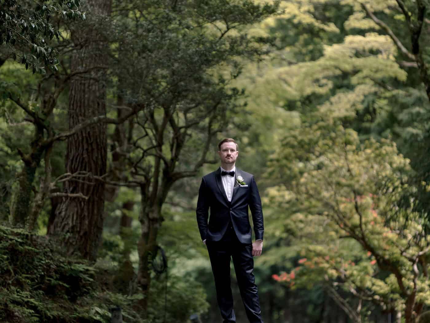 The groom stands alone in black tie, a white rose at his lapel, among tall trees and ferns on the day of his forest wedding in Japan.