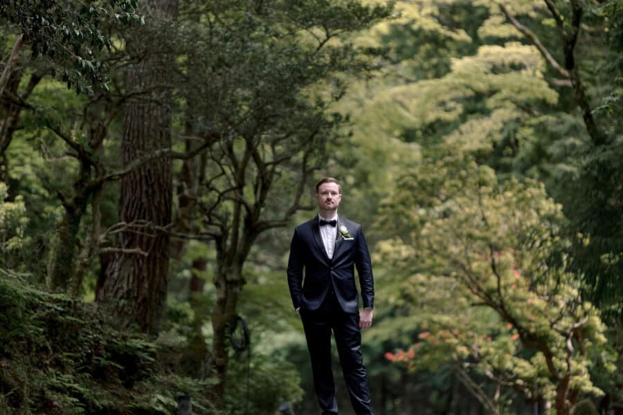 The groom stands alone in black tie, a white rose at his lapel, among tall trees and ferns on the day of his forest wedding in Japan.