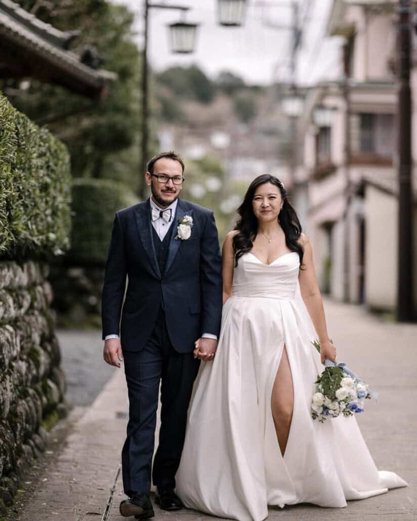 The bride and groom walk hand in hand down a quiet Shuzenji Onsen street, past a stone wall, clipped hedges and old street lamps.