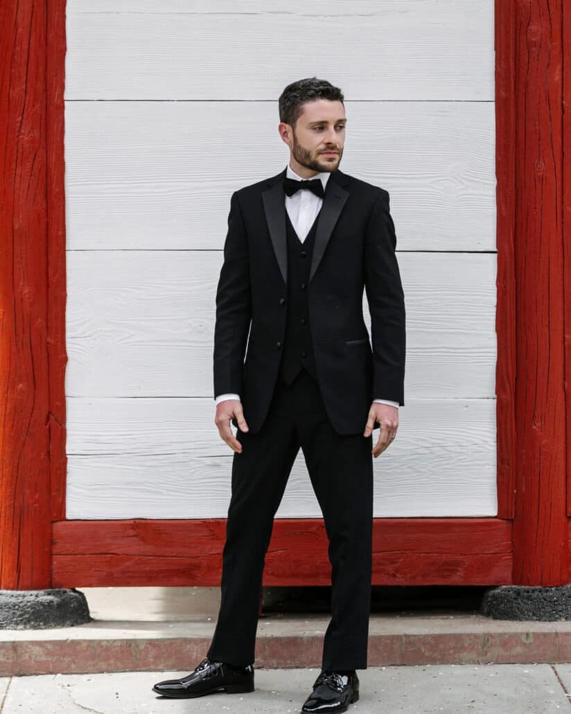 The groom in a black three-piece tuxedo, bow tie and patent shoes stands against a red-and-white wall at a Shinto shrine near Lake Kawaguchi.