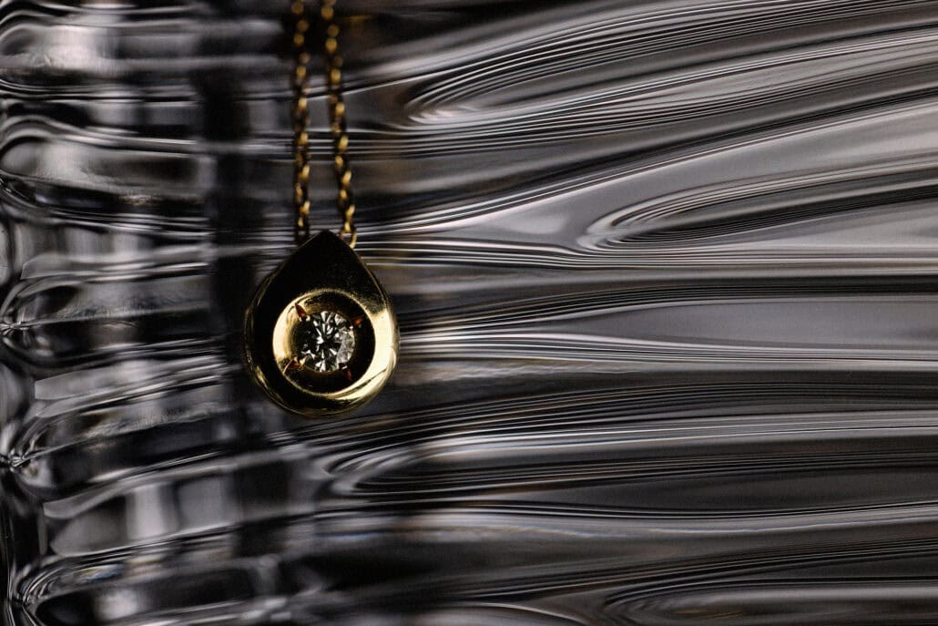 A gold teardrop pendant set with a single clear stone hangs on a fine chain against fluted glass, on the morning of a wedding in Okinawa.