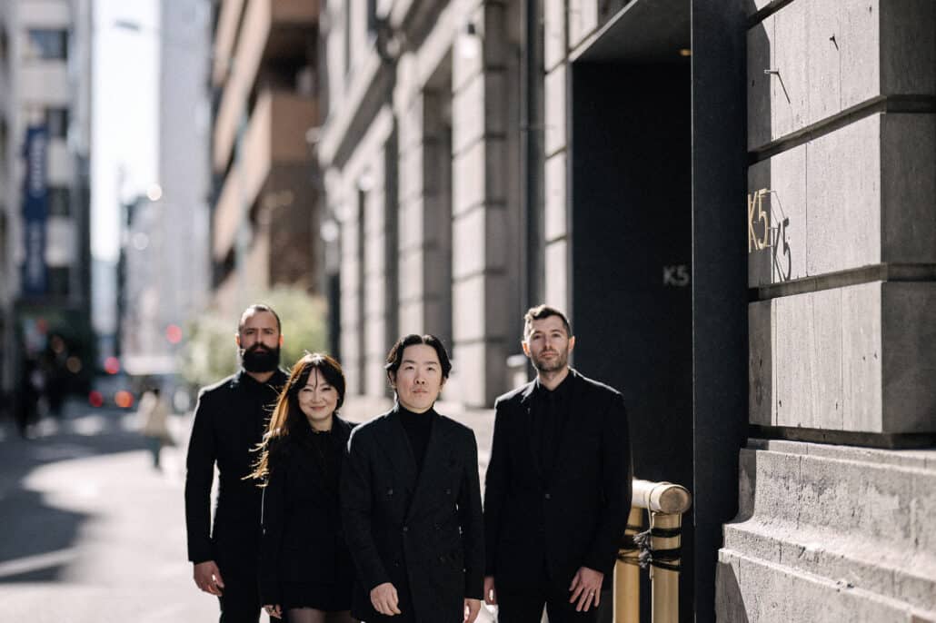 The groom and his grooms people, all in black, stand on a sunlit street in Kabutocho beside the K5 letters on the stone face of the old bank building.