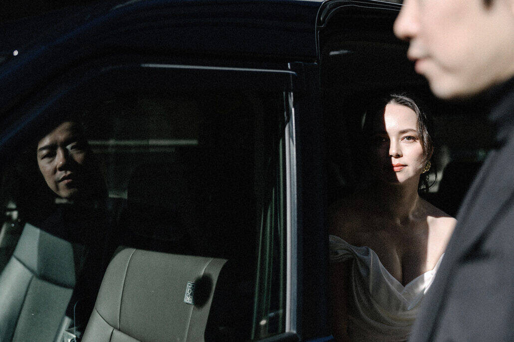 The bride sits in a slice of sun in a Tokyo taxi doorway, the groom reflected in the dark window and his profile blurred in the foreground.