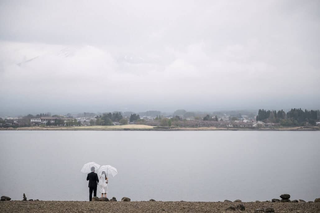 Seen from behind, the bride and groom stand under two clear umbrellas on the stony shore of Lake Kawaguchi, Mt Fuji faint in the cloud above.