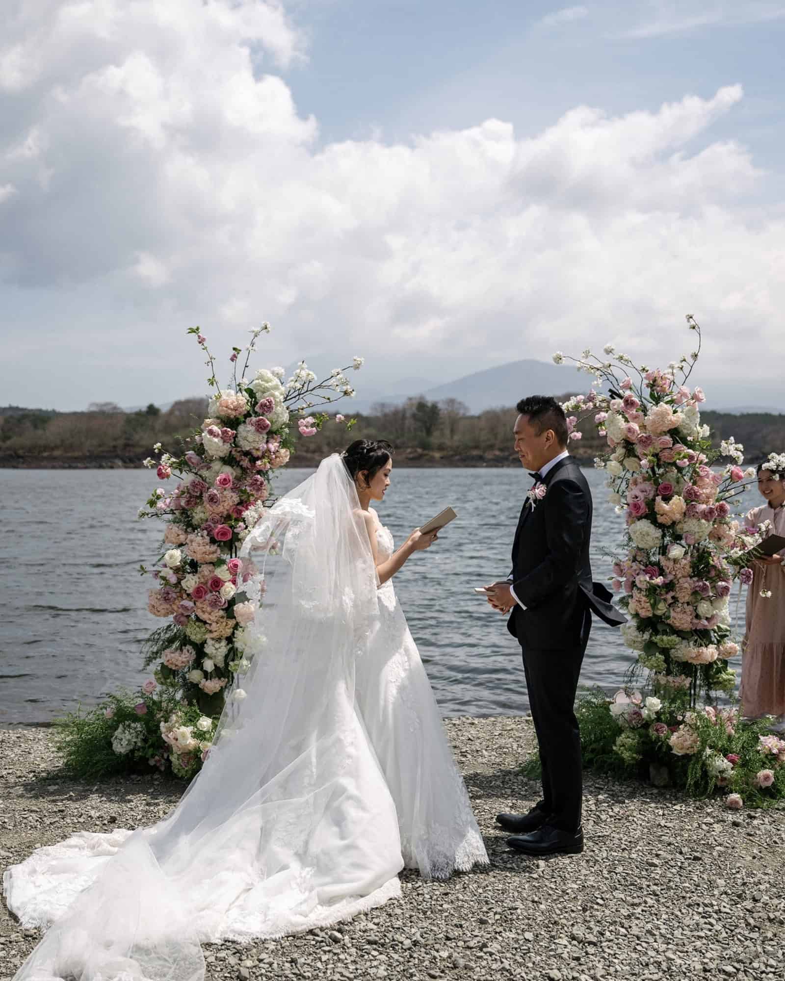 The bride reads her vows from a small book between two floral columns on the shore of Lake Shoji, an outdoor wedding venue near Mt Fuji.