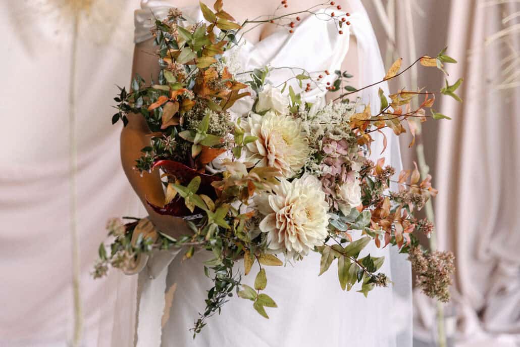 The bride's bouquet in autumn colours up close, with cream dahlias, pale roses, pink hydrangea, rose hips and turning leaves against her satin gown.