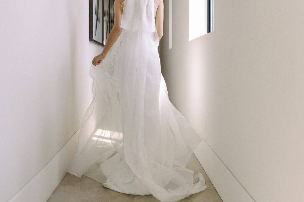The bride walks down a bright white corridor at Rosewood Miyakojima, the long sheer train of her halter overdress spread across the pale floor.