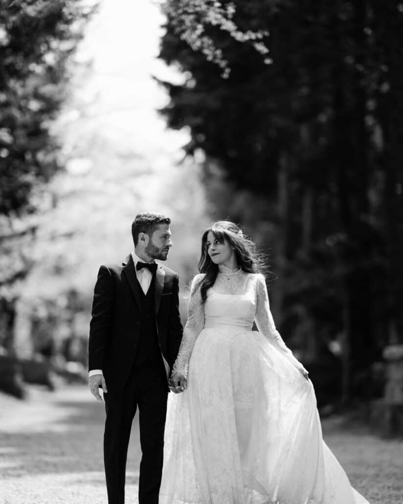 In black and white, the couple walk hand in hand along a cedar-lined shrine approach near Lake Kawaguchi, past a stone lantern.