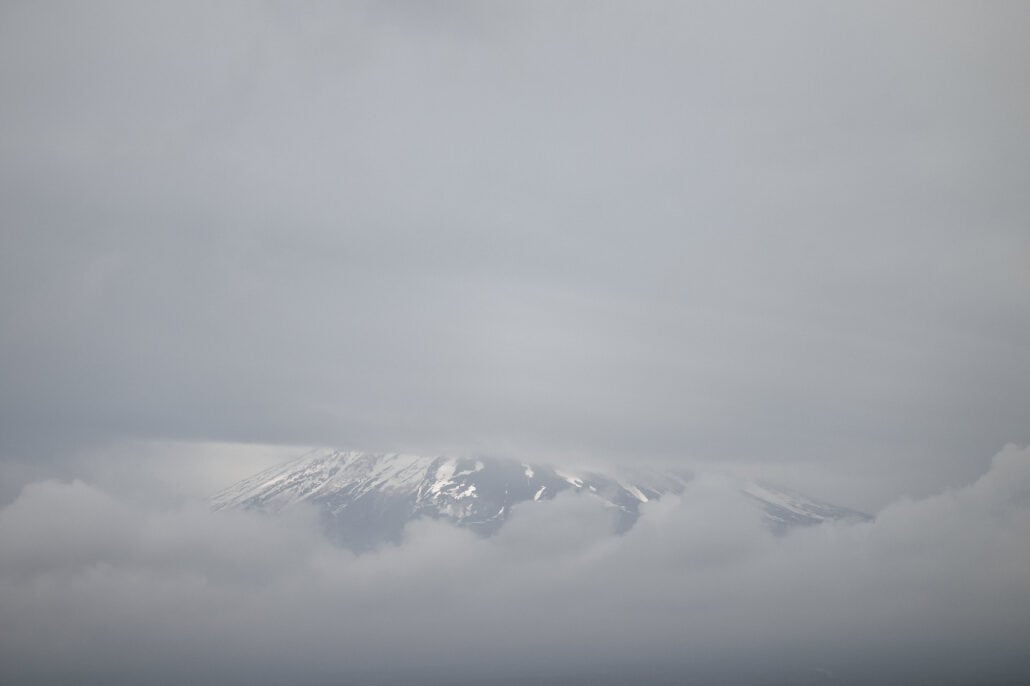 The snow streaked upper slopes of Mt Fuji appear through a narrow gap between layers of heavy grey cloud on a spring morning.