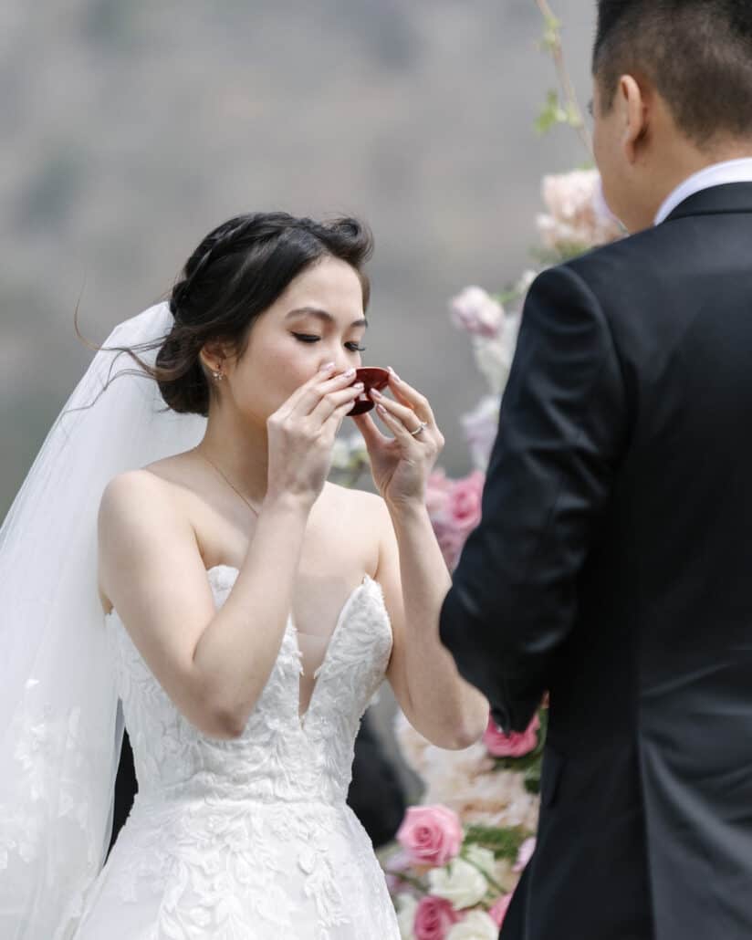 The bride sips sake from a red lacquer cup in san san kudo, the Japanese sake ceremony, as the groom faces her at their Lake Shoji ceremony.