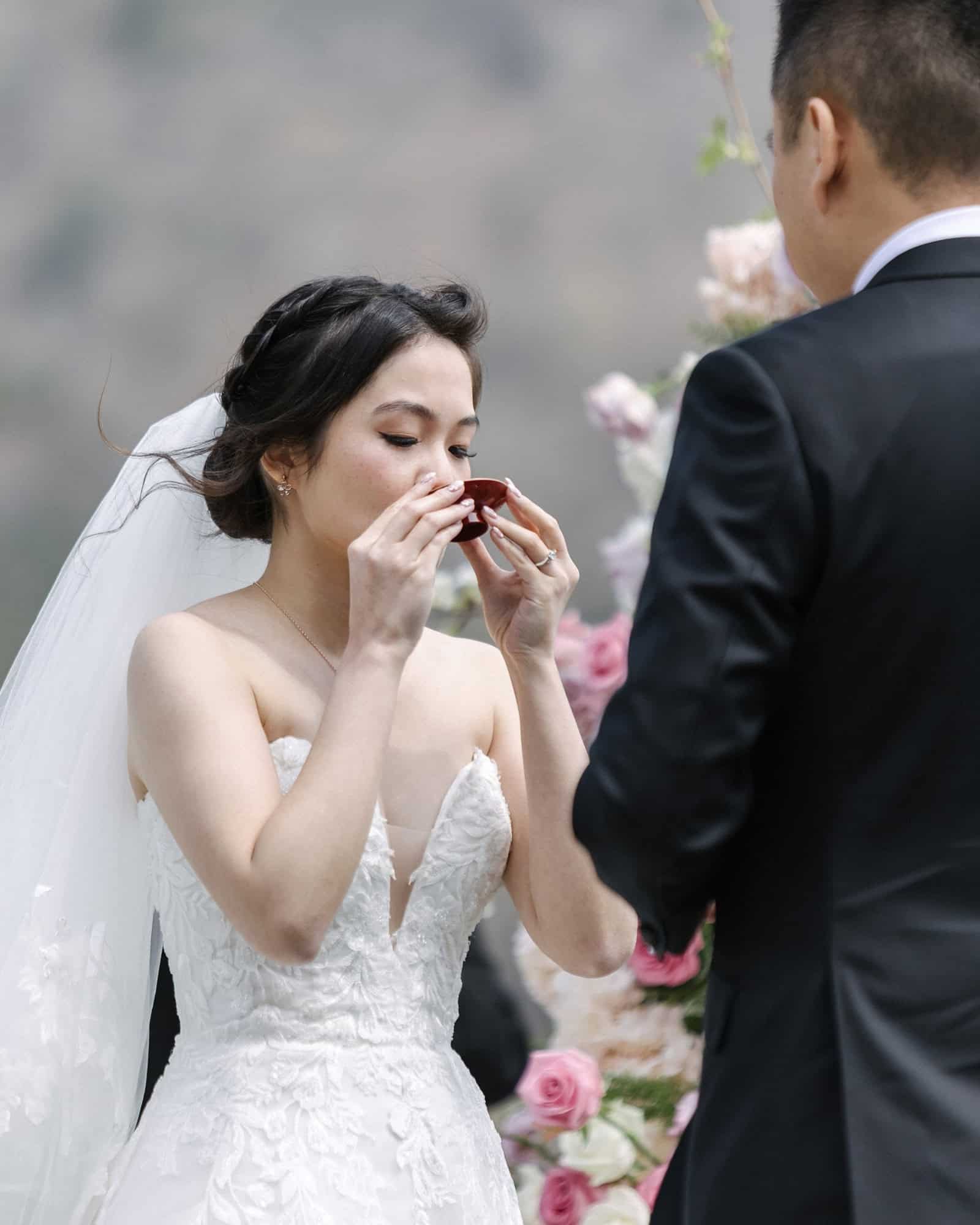 The bride sips sake from a red lacquer cup as the groom faces her during san-san-kudo, the Japanese sake ceremony, at a Lake Shoji wedding.