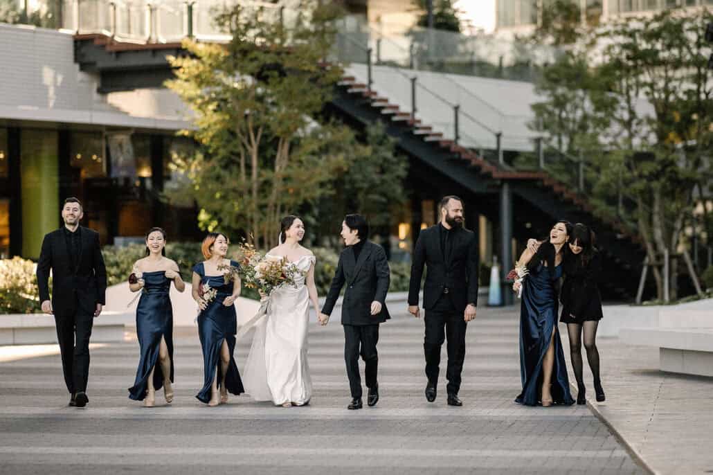 The couple walk hand in hand across a plaza in Takeshiba with bridesmaids in navy satin and grooms people in black, laughing before the ceremony.