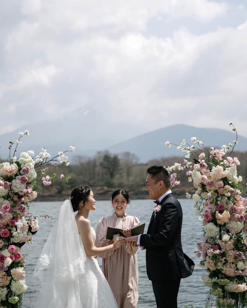 The officiant laughs between the bride and groom at a floral arch of pink roses and blossom, with Lake Shoji and the hills behind them.