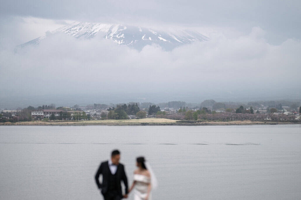 Mt Fuji's snowy slopes stand sharp above the cloud across Lake Kawaguchi, the bride and groom a soft blur walking hand in hand in front.