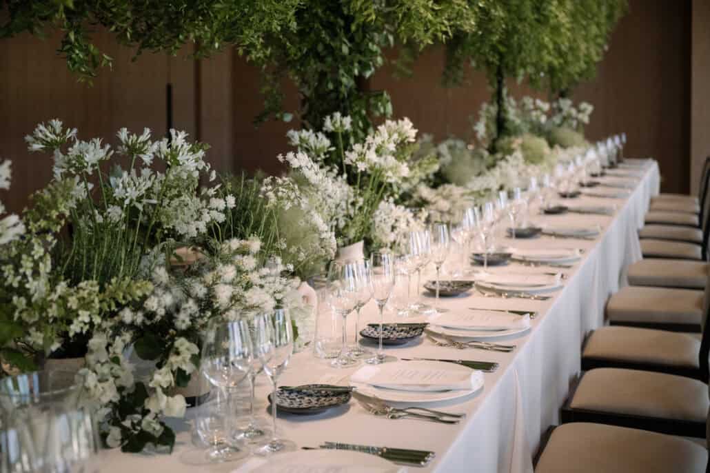 One long table set for the wedding lunch at Rosewood Miyakojima, white flowers and glassware down its length and greenery hanging from the ceiling.