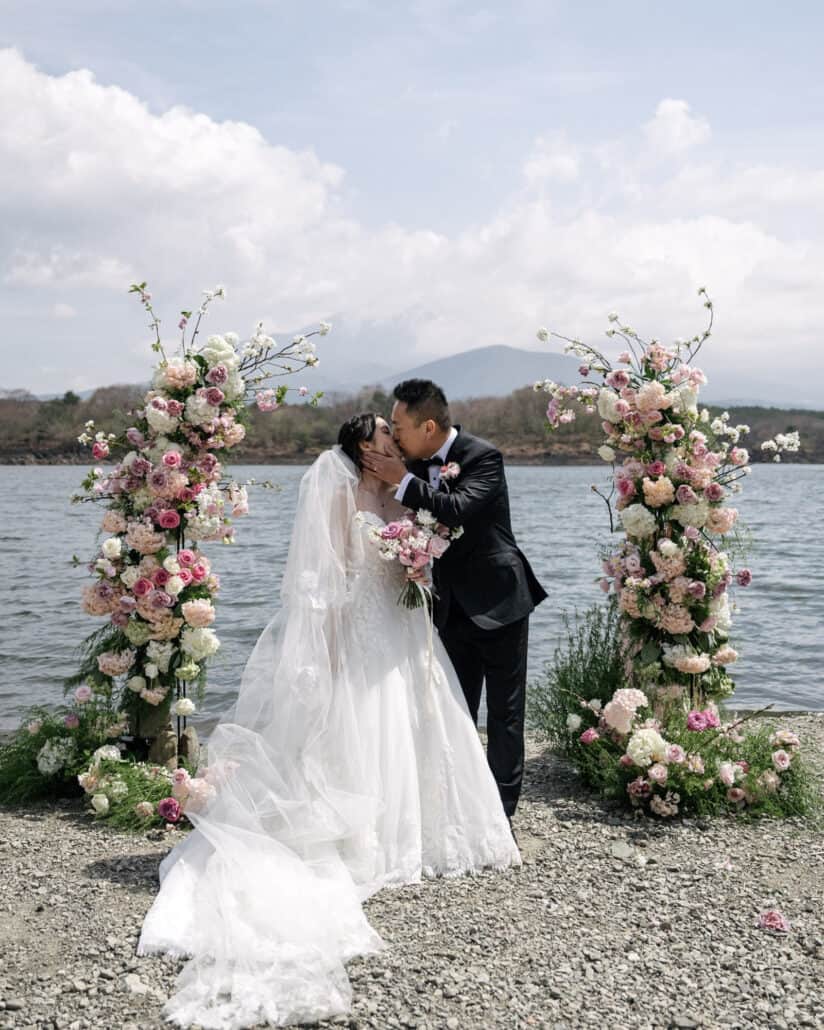 The bride and groom share their first kiss between two floral columns at a Lake Shoji wedding ceremony, the water and a mountain behind them.