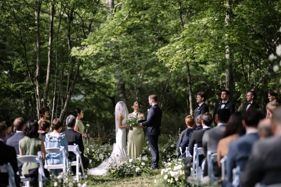 The groom holds the veiled bride's hands as the officiant speaks at a forest wedding in Japan, guests on white chairs and the wedding party beside.