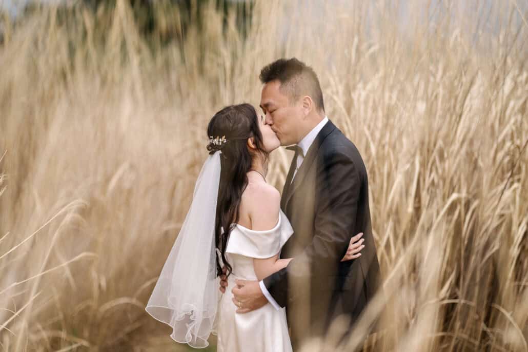 The bride and groom kiss in tall golden grass, his arms around her waist, her short veil falling from a pearl hair pin.