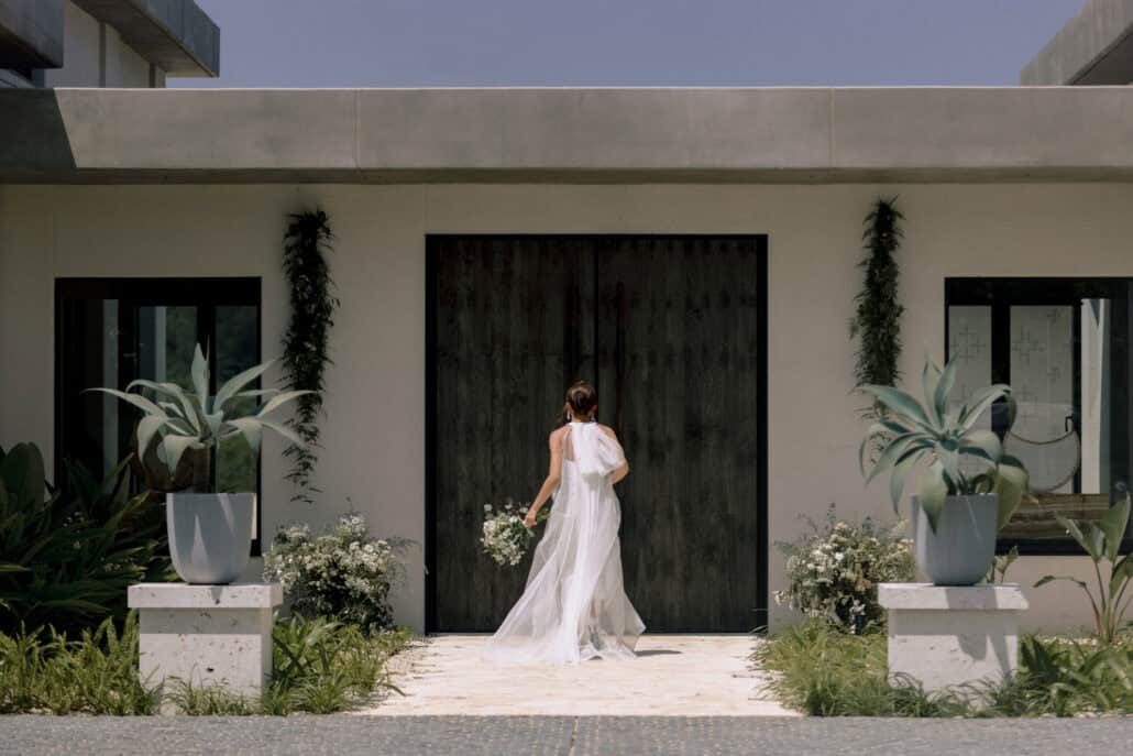The bride walks toward tall black timber doors with her bouquet, a large bow at the back of her dress and agaves either side, at Rosewood Miyakojima.