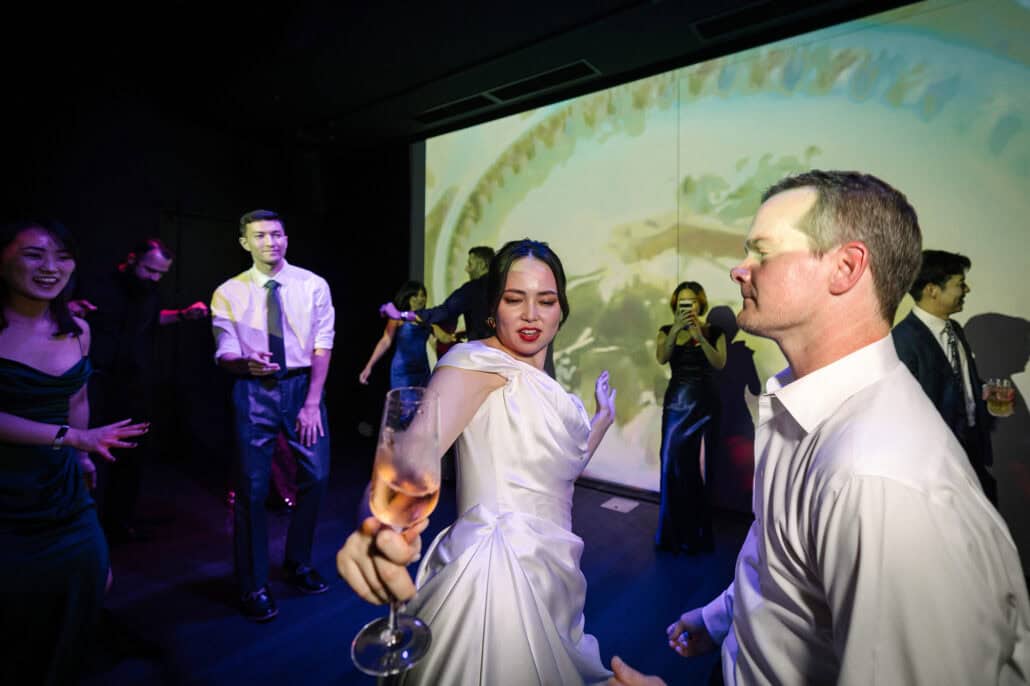 The bride dances with a glass of rosé among friends in front of the projection wall at BANK30 as a bridesmaid films, the party in full swing.
