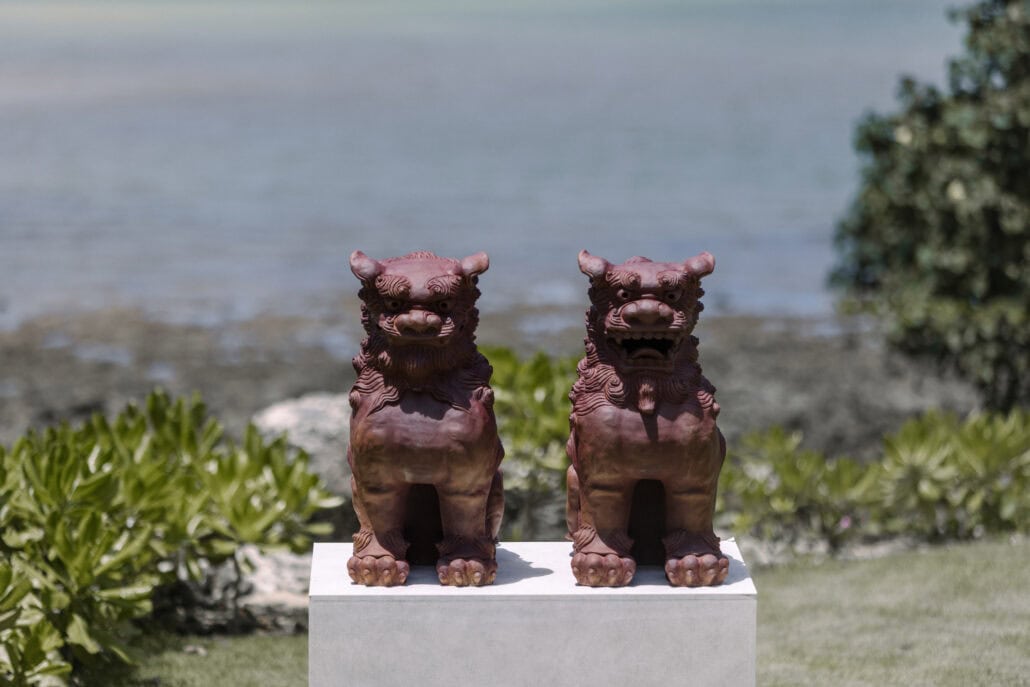 A pair of red clay Okinawa shisa, one mouth closed and one open, stand on a white plinth above the reef and the sea at a Miyakojima wedding.