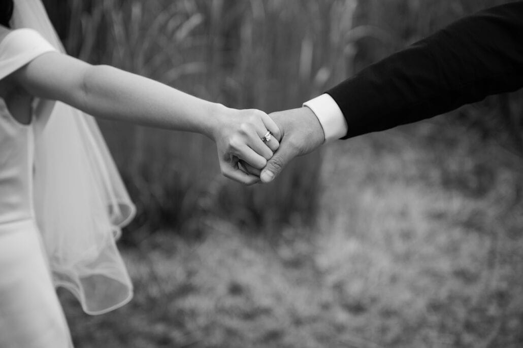 In black and white, the bride and groom hold hands at arm's length, her wedding band and solitaire ring showing beside his white cuff and dark sleeve.