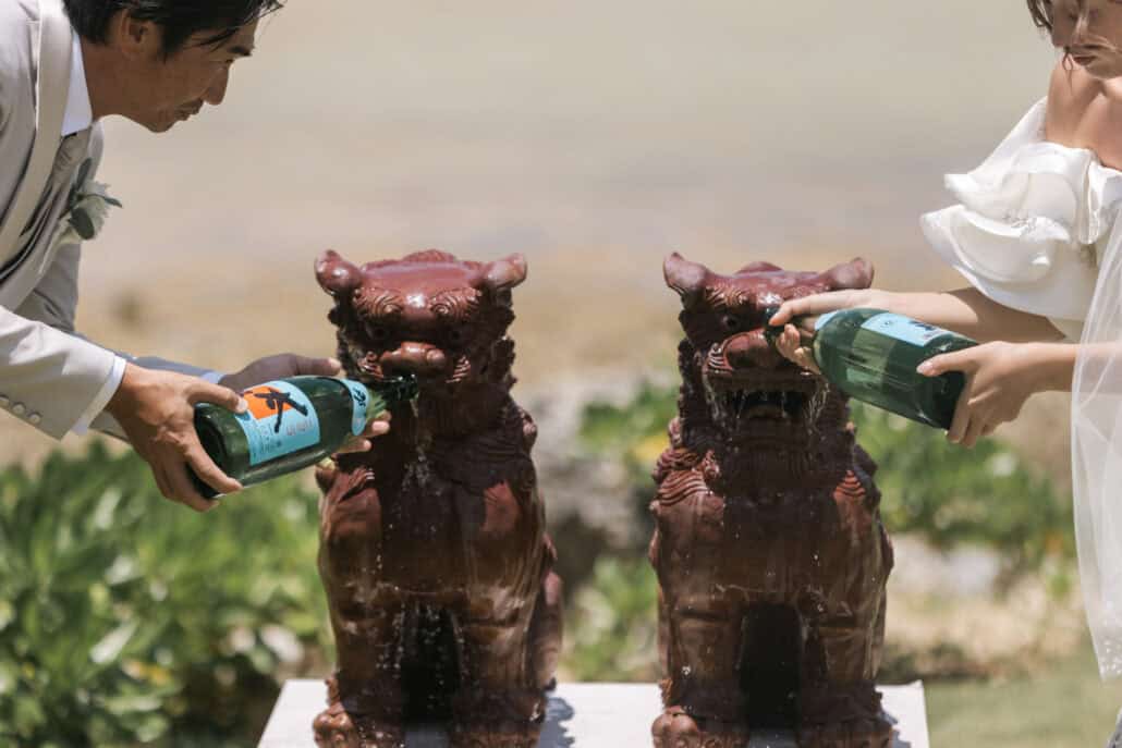 The bride and groom pour from green glass bottles over the heads of a pair of red clay shisa before their Rosewood Miyakojima wedding ceremony.