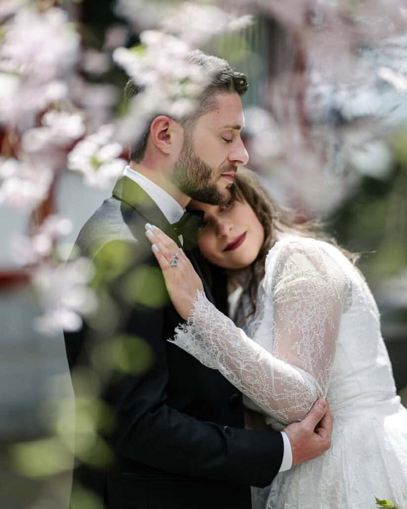 The bride rests her head under the groom's chin, his eyes closed, her blue-stone ring on his chest, through cherry blossom at a Japanese shrine.