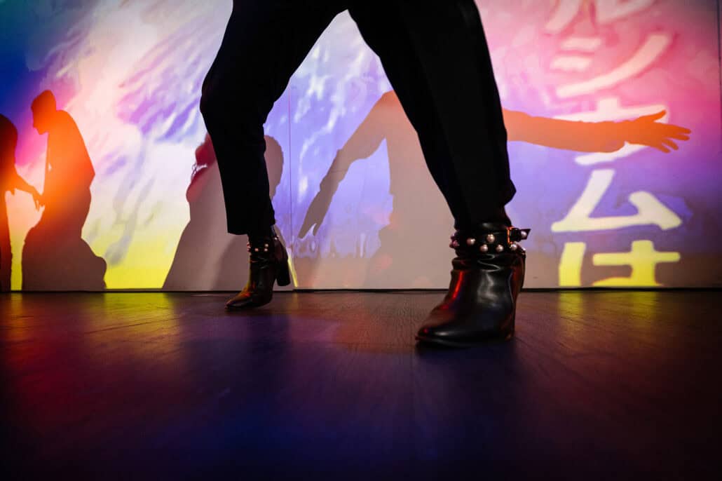 Studded black ankle boots mid step on the dance floor, with dancers' shadows and Japanese text projected behind at a wedding party in Tokyo.